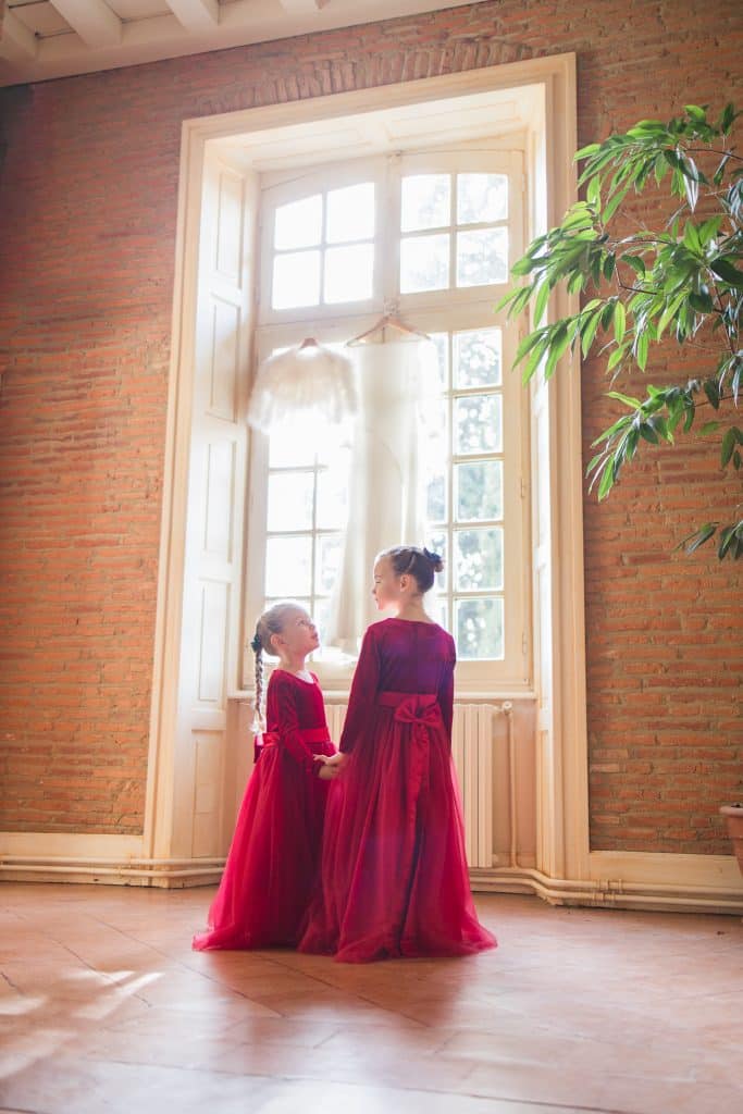 deux jeunes filles habill&eacute;es en robes rouges sont devant la fenetre sur laquelle il y a une robe de mari&eacute;e