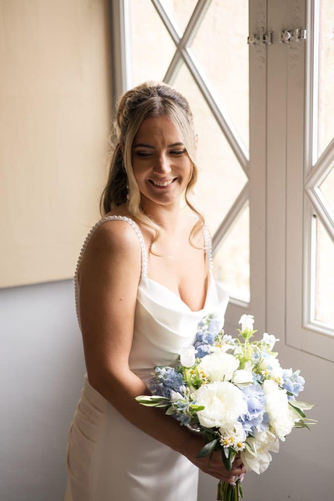 Une femme en robe de mari&eacute;e avec un bouquet des fleurs blanches et bleues devant une fenetre
