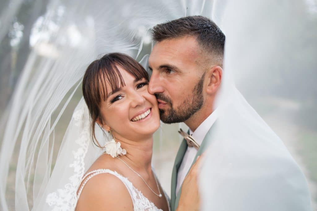 un couple de mari&eacute; qui posent sous le voile le mari&eacute; fait un bious &agrave; la joue de la mari&eacute;e